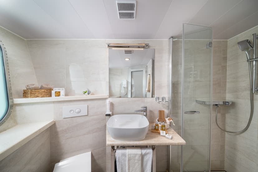 Modern cabin bathroom with vessel sink, lighted mirror, and glass shower stall; porthole window visible.