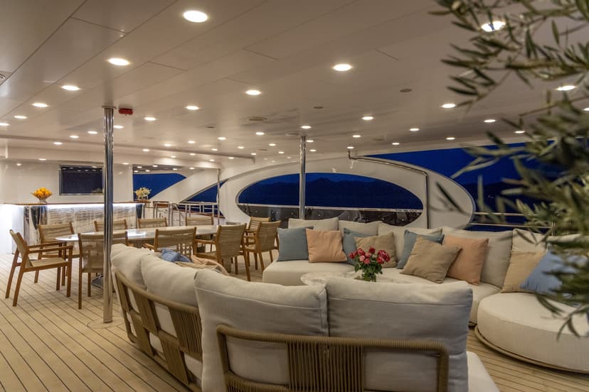 Luxury yacht deck seating area with dining tables and dark blue night view of mountains.