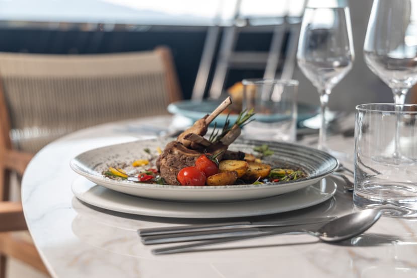 Rack of lamb with roasted potatoes and tomatoes served on a marble table with wine glasses.