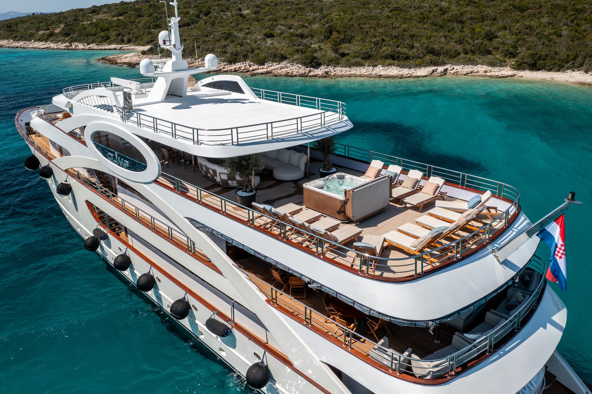Luxury yacht Olimp anchored in turquoise coastal waters near a green, rocky shore with Croatian flag.
