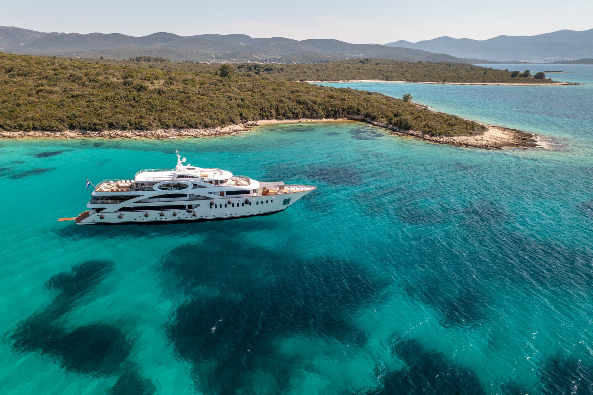 Luxury yacht Olimp anchored in turquoise coastal waters near a wooded island and hills.