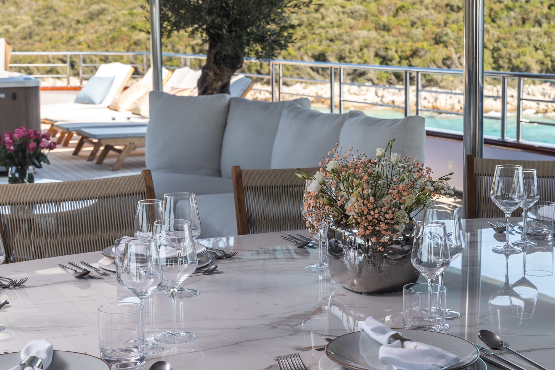 Outdoor dining area with set marble table, white cushions, and view of green hillside and turquoise water.