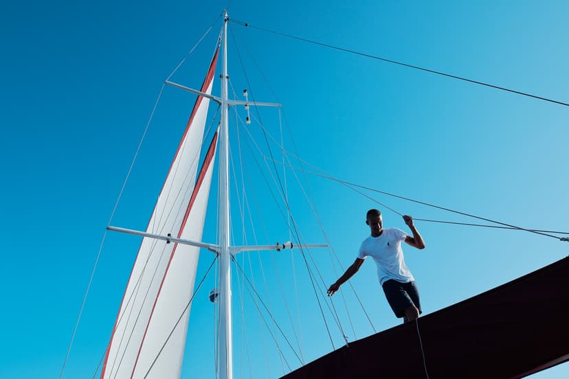 Sailor managing lines near the mast and white sail against a clear blue sky.