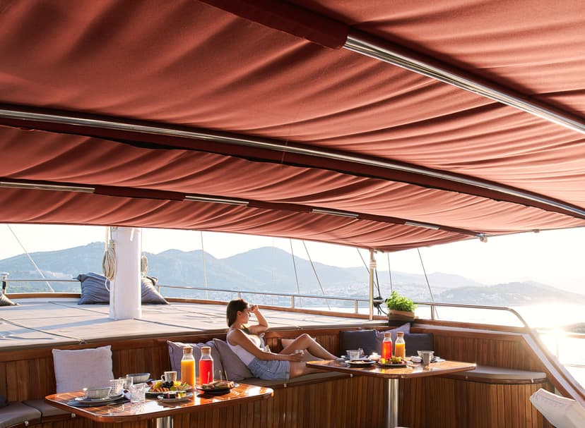 Woman relaxing with breakfast on wooden yacht deck overlooking coastal mountains at sunrise.
