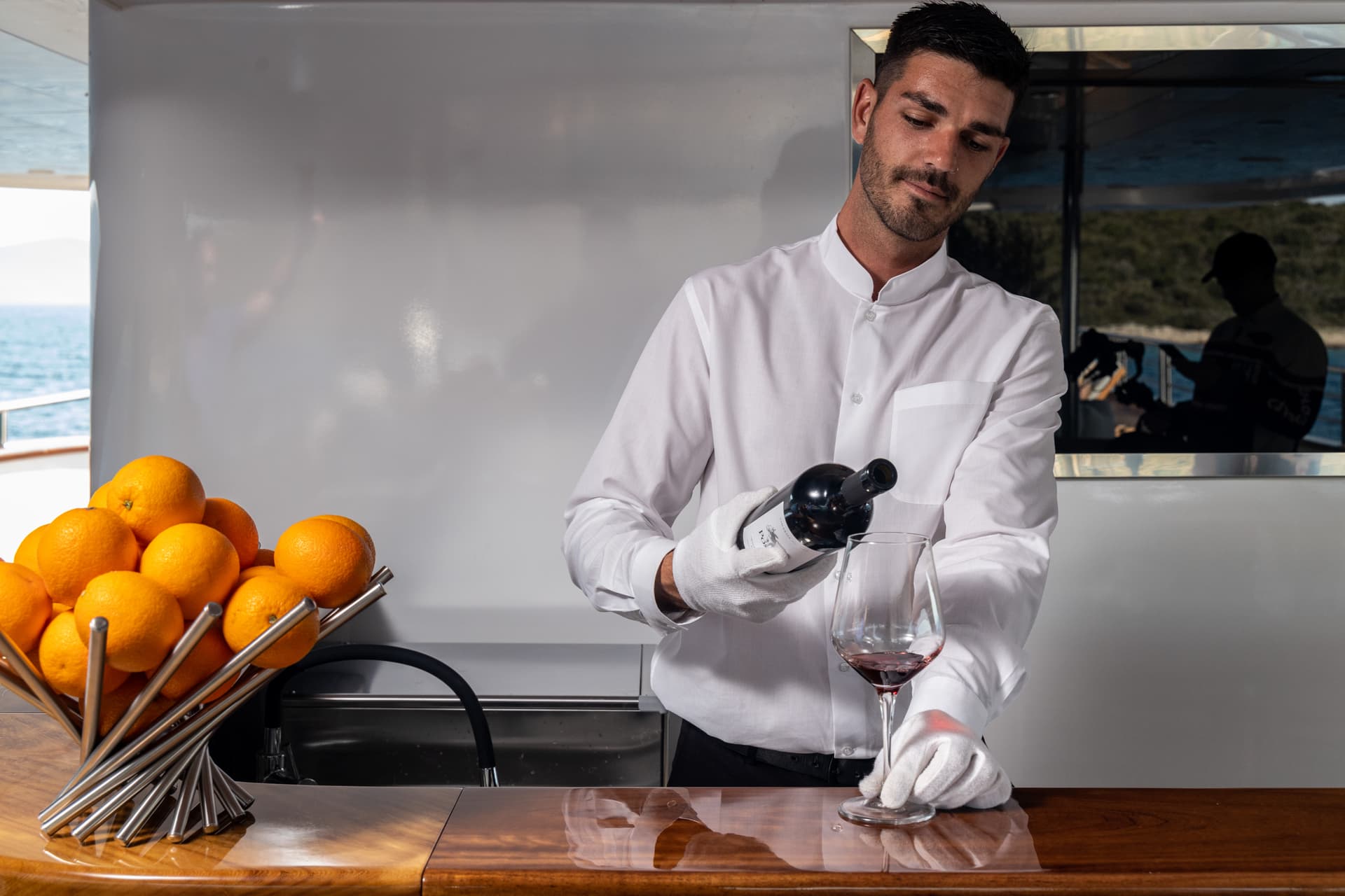 Bartender in white shirt pouring red wine on yacht with sea view and oranges.