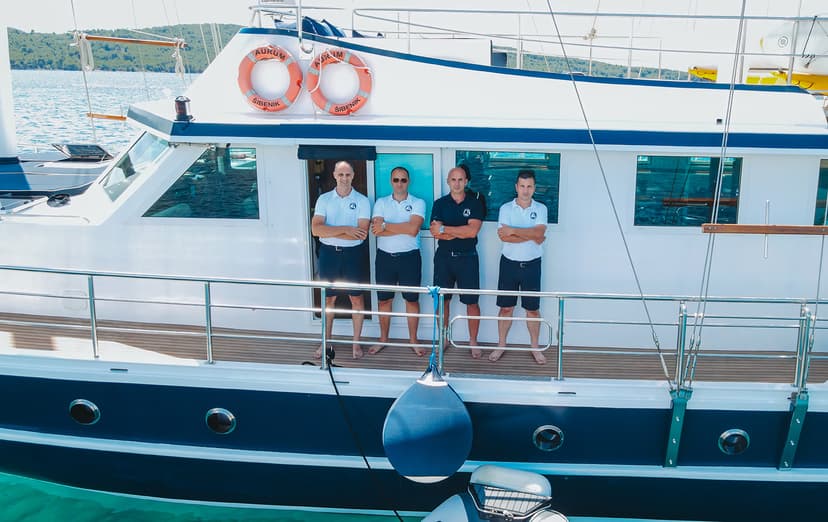 Four crew members standing on a white and blue yacht with "Šibenik" life rings in clear coastal water.