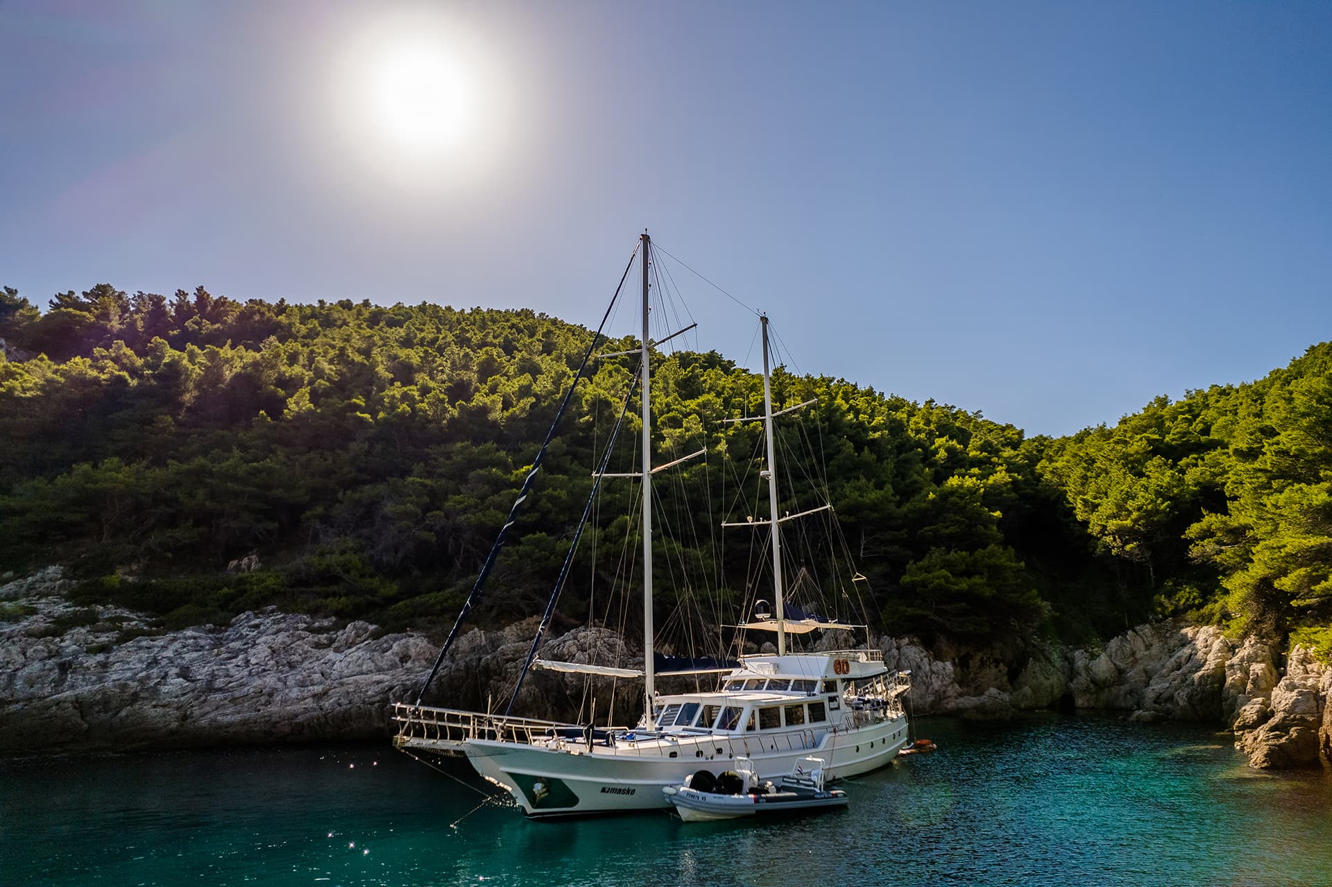 Gulet sailboat anchored in turquoise cove with rocky, forested coastline under bright sun.