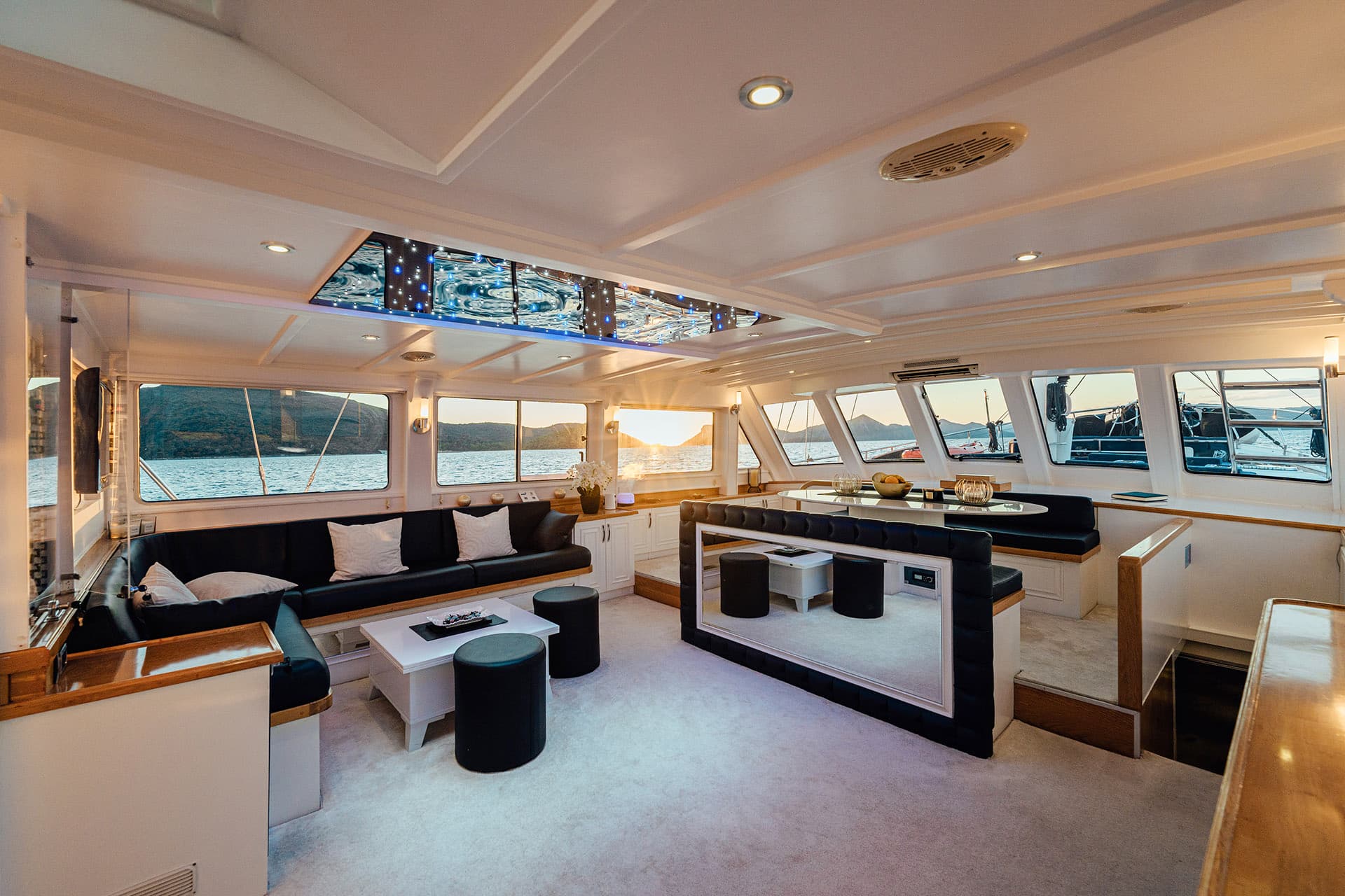 Cosy gulet interior with black seating, white decor, and sunset view over water and hills.