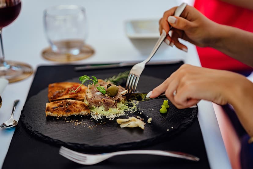 Hands cutting meat tartare with avocado and toast on a black slate plate at a table.