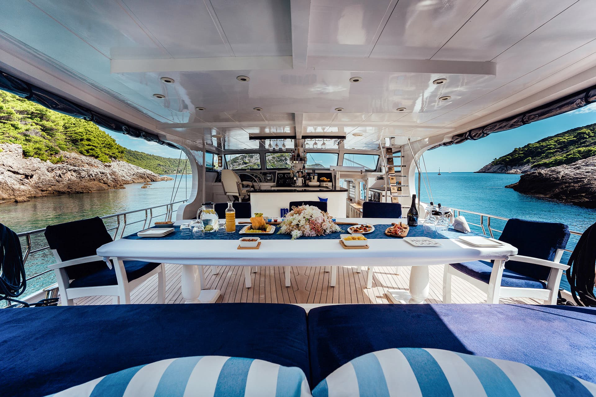 Dining table set on a gulet sundeck anchored near a rocky, green coastline.
