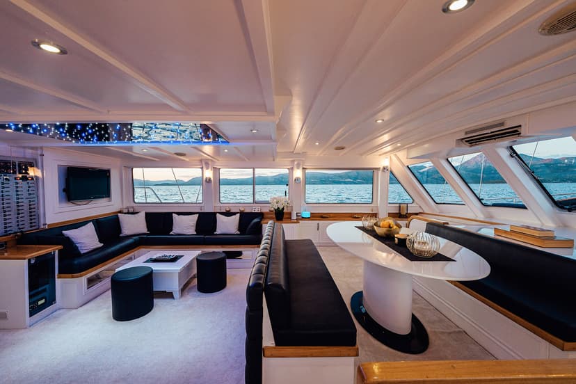 Interior of a yacht salon with black seating, white tables, and mountain views on the water.