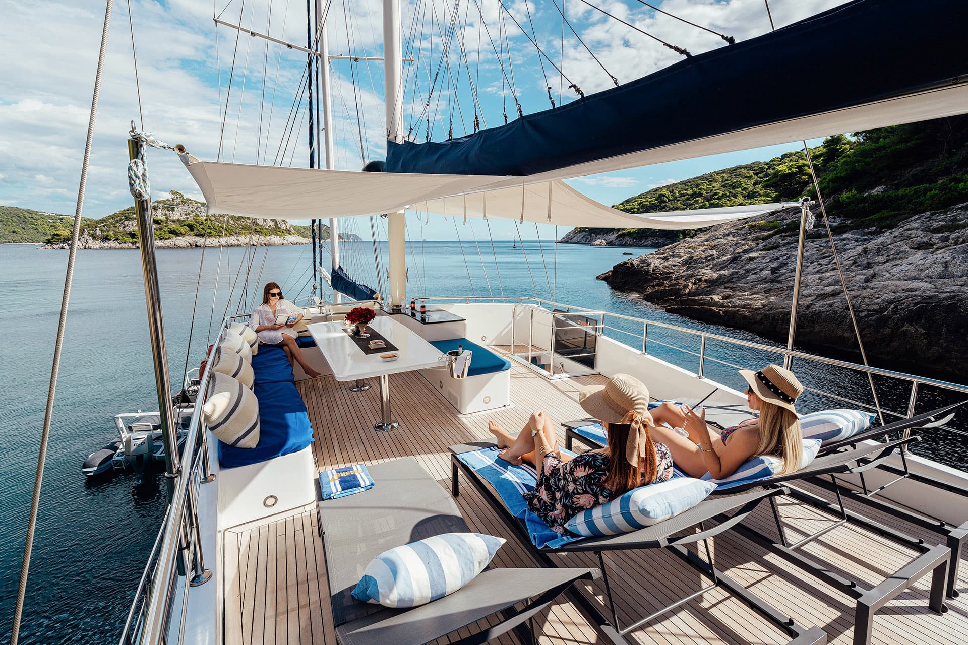 Sunbathing on a Gulet deck near a rocky, green coastline under a blue sky.