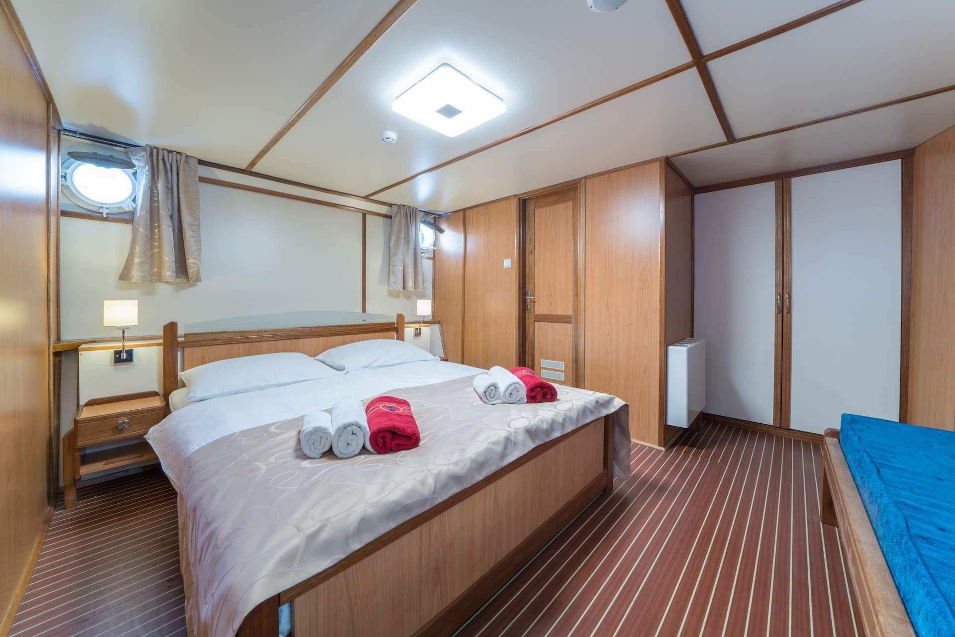 Cabin bedroom with wood paneling, porthole window, and neatly folded towels on the bed.