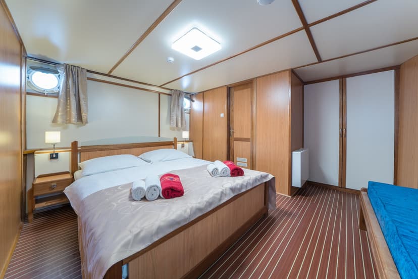Cabin bedroom with wood paneling, porthole window, and neatly folded towels on the bed.
