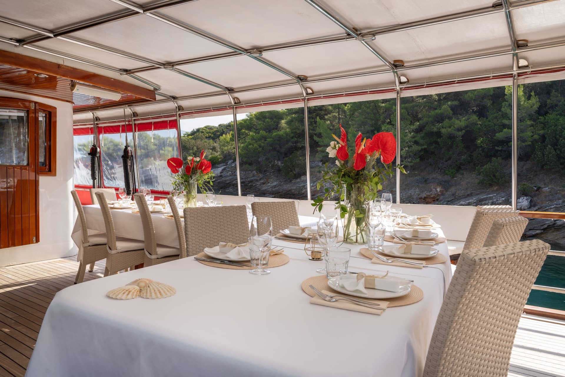 Dining table set for a meal on a gulet deck near a wooded coastline.