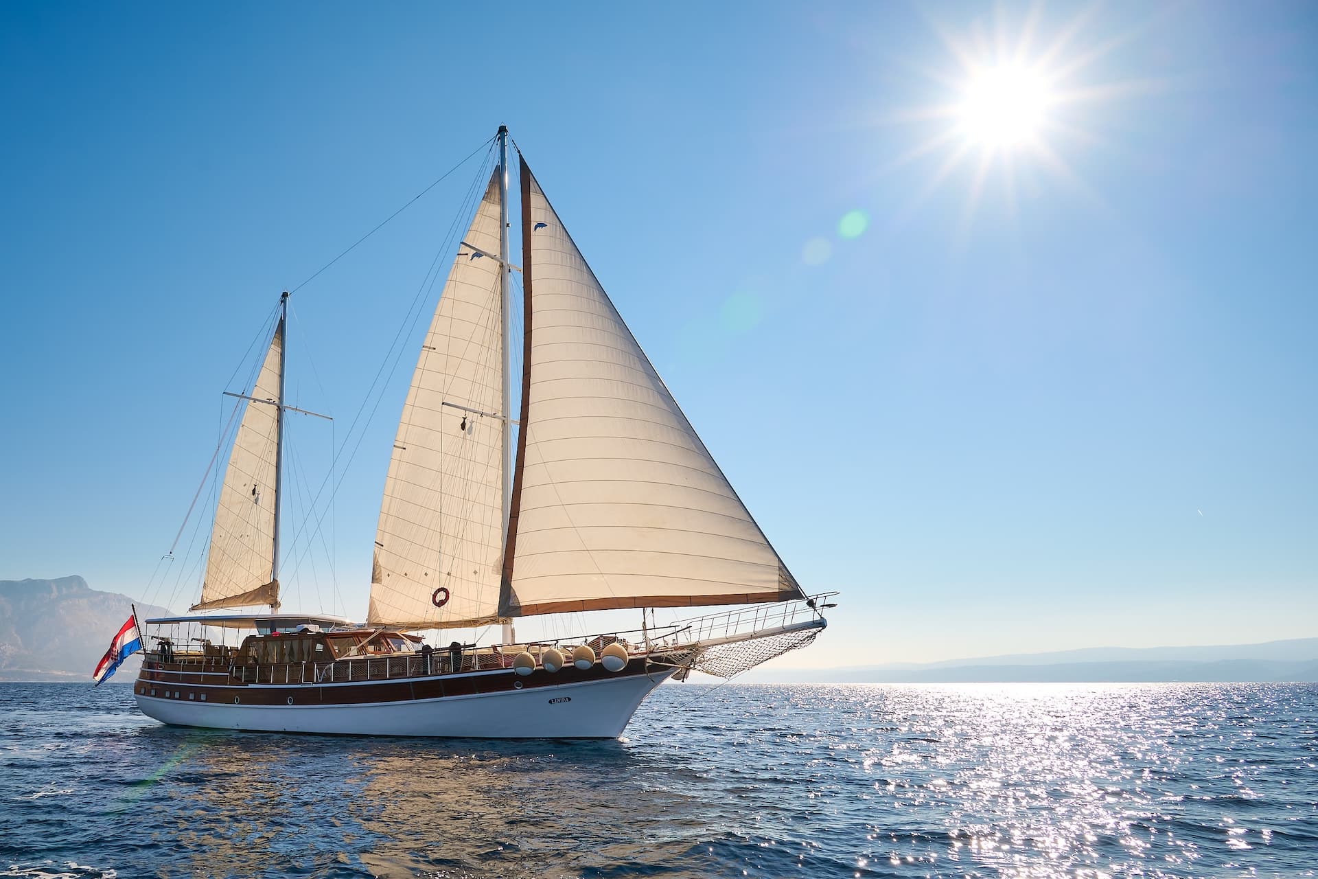 Sailboat named Linda with white sails on sparkling blue sea near mountains under bright sun.