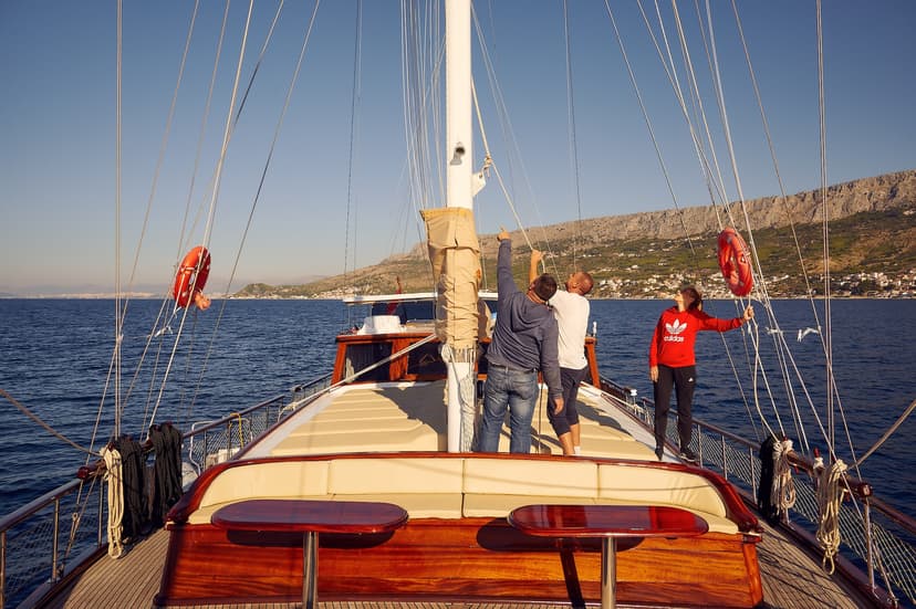 People on a wooden sailboat adjusting rigging near a rocky, coastal town under clear blue sky.