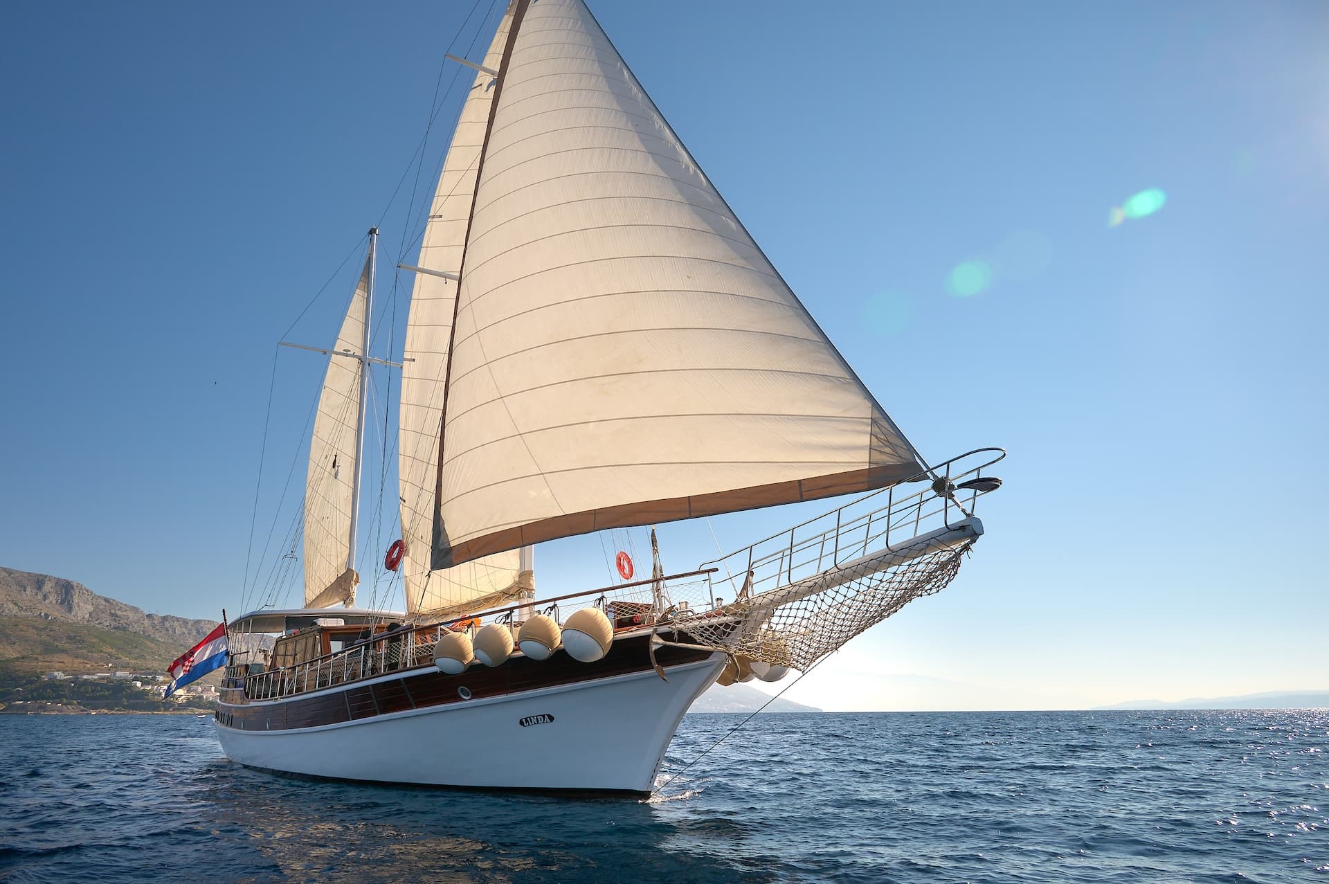Sailboat named Linda with sails up on blue sea near rocky coastline flying Croatian flag