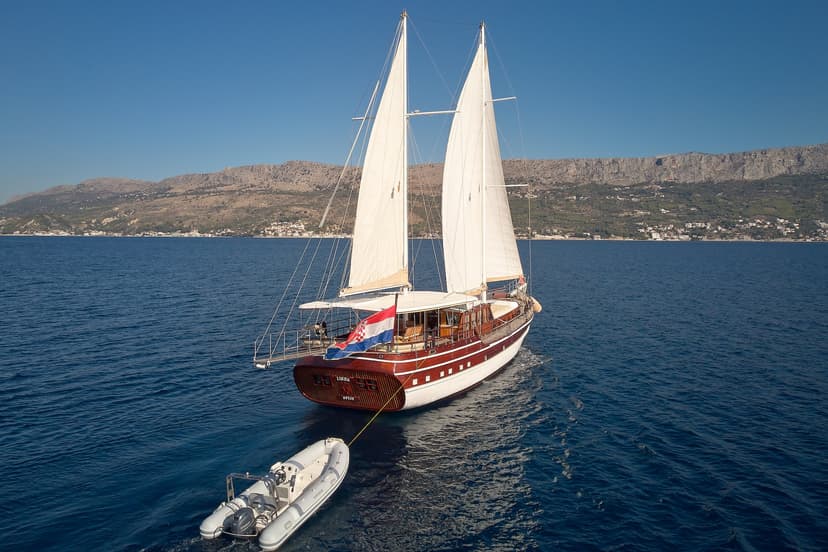 Wooden sailboat with sails up towing a dinghy near the Croatian coastline.