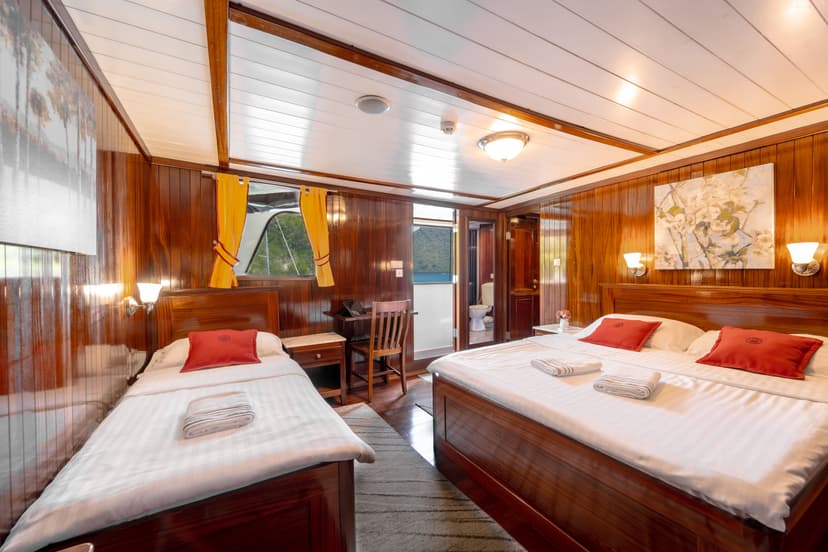 Cabin interior with double and single beds, wood paneling, and view of green hills over water.