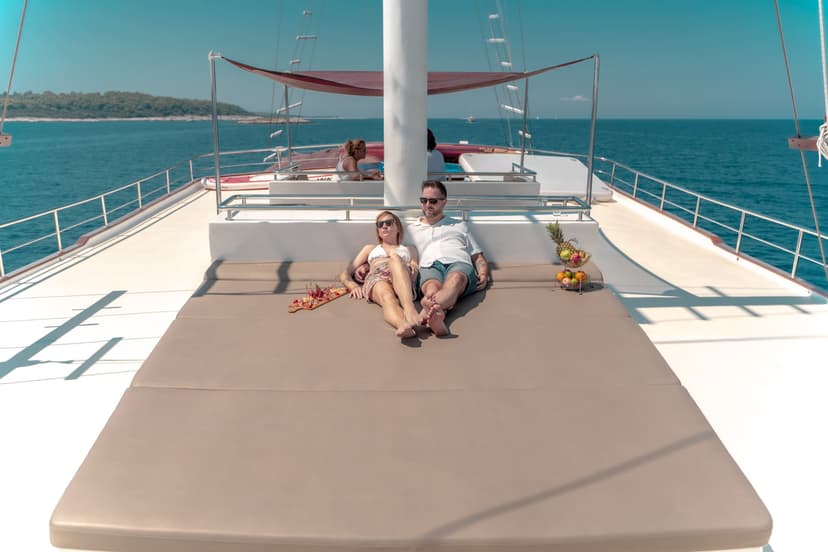 Couple relaxing on sun deck of yacht with fruit platter and charcuterie board, blue sea background.