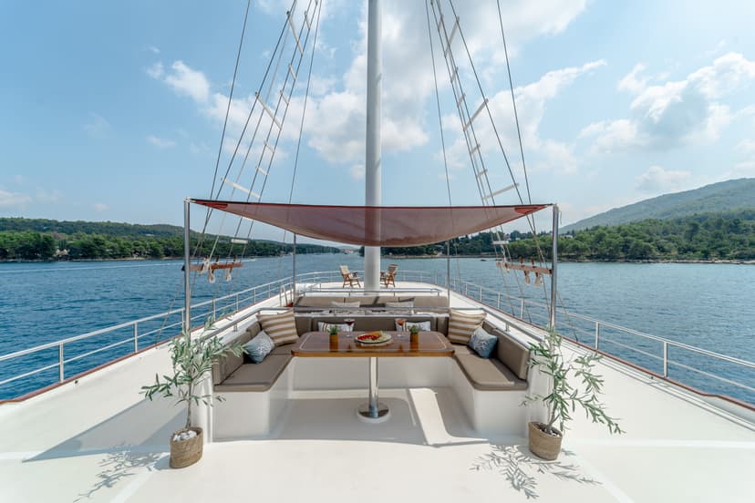 Bow seating area of a yacht with dining table, shaded by a canopy, cruising near a forested coastline.