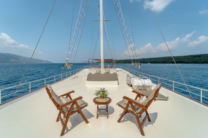 Deck of a boat with wooden chairs, table, and rigging sailing past forested coastline.