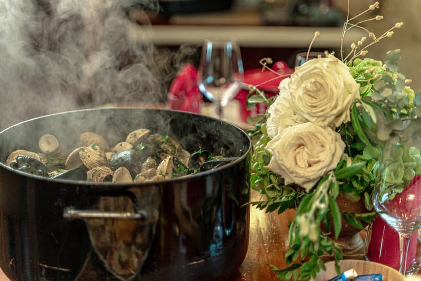 Steaming pot of mussels and clams cooking, next to white roses centerpiece on wooden table.