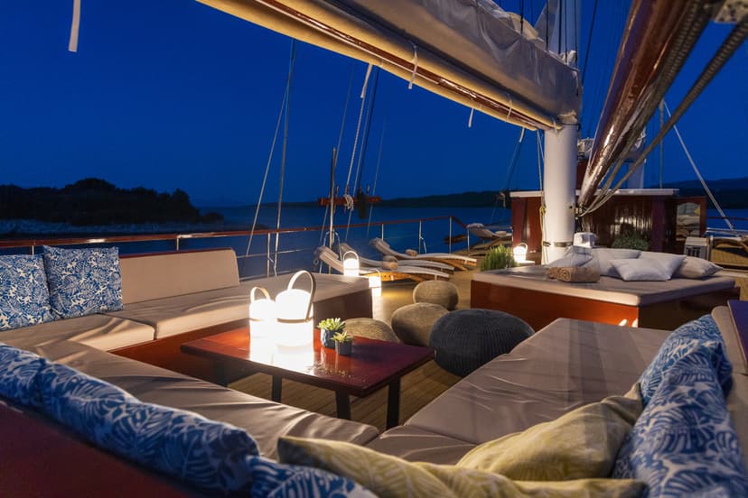 Yacht deck lounge area with seating and lanterns at night near a dark coastline