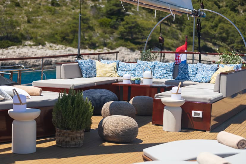 Yacht deck lounge with cushions and poufs, anchored near a green, rocky coastline.