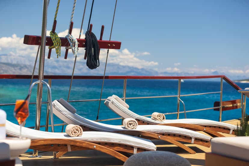Sun loungers on a boat deck with ropes hanging, overlooking turquoise sea and distant mountains.