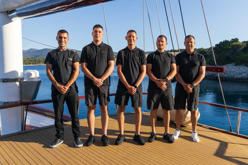 Five crew members standing on a wooden boat deck near a forested coastline.