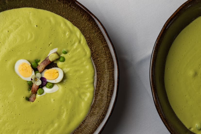 Green cream soup garnished with halved eggs, bacon, and peas in a brown bowl.