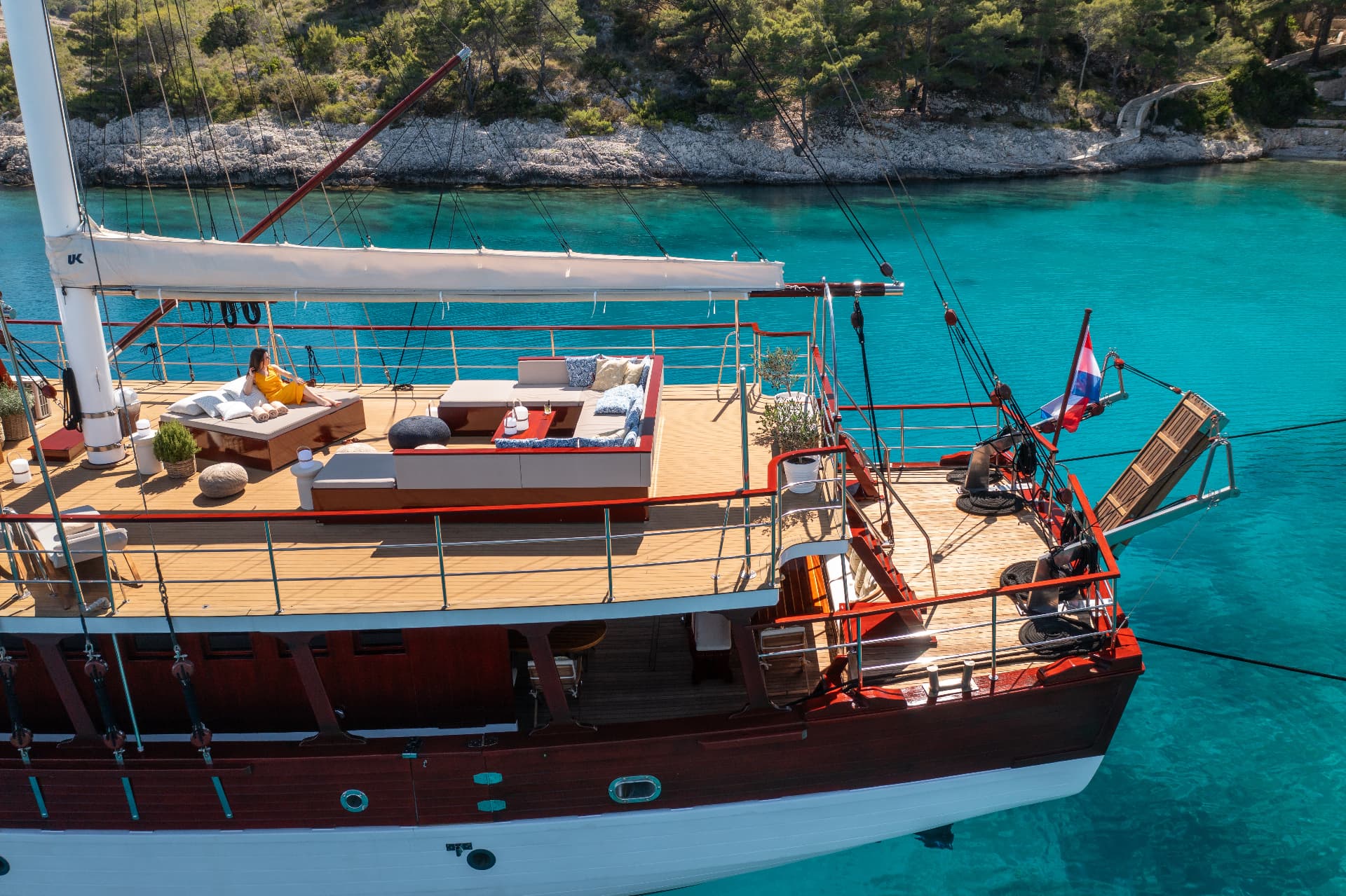 Sailboat with wooden deck anchored in turquoise coastal waters near a rocky, forested shore.