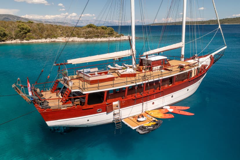 Large wooden sailboat anchored in turquoise water near a wooded island, Croatian flag visible.