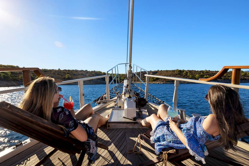 Two women relax on wooden deck chairs drinking cocktails on a boat near a wooded coastline.