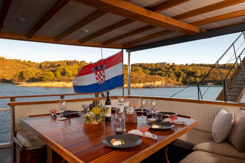 Outdoor dining table set on boat deck with Croatian flag near wooded coastline.