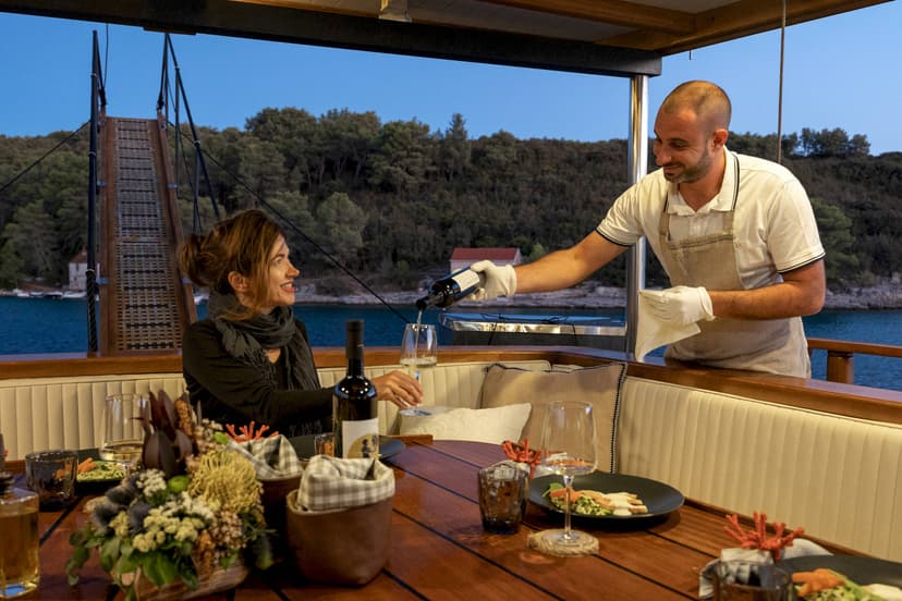 Dining on a boat deck as crew pours wine near a forested coastline at dusk.