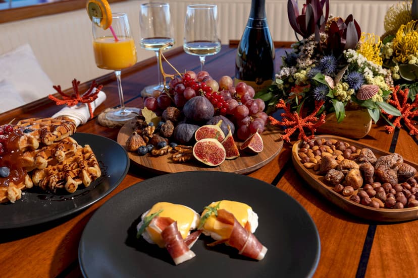 Brunch spread with waffles, eggs benedict, fruit, nuts, and champagne on a wooden deck.