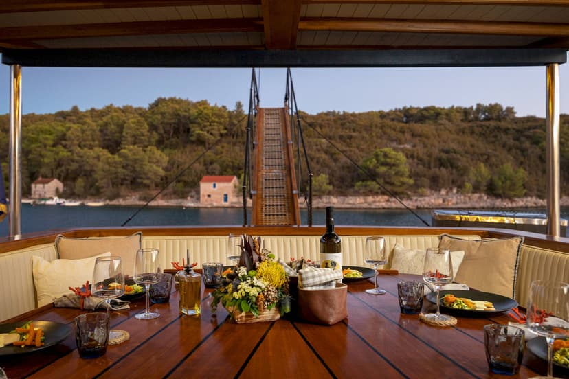 Dining setup on a boat deck overlooking a wooded coastline and gangplank.