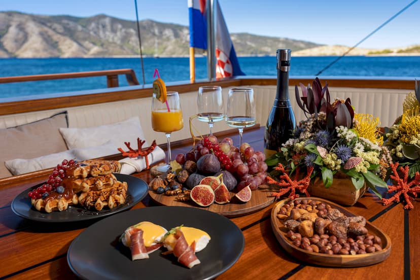 Brunch spread with waffles and eggs benedict on a boat deck overlooking mountains and blue sea.