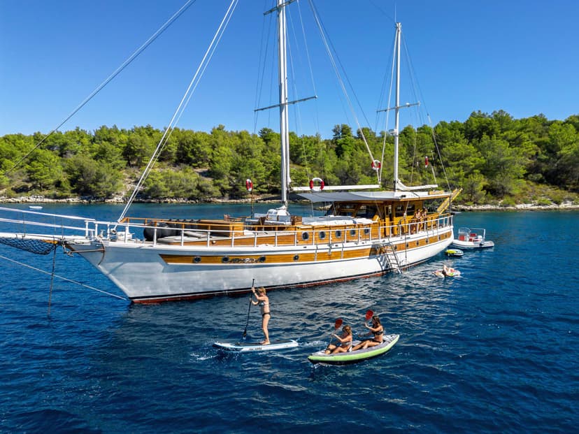 Large wooden sailboat anchored near forested coastline with people paddleboarding and kayaking.