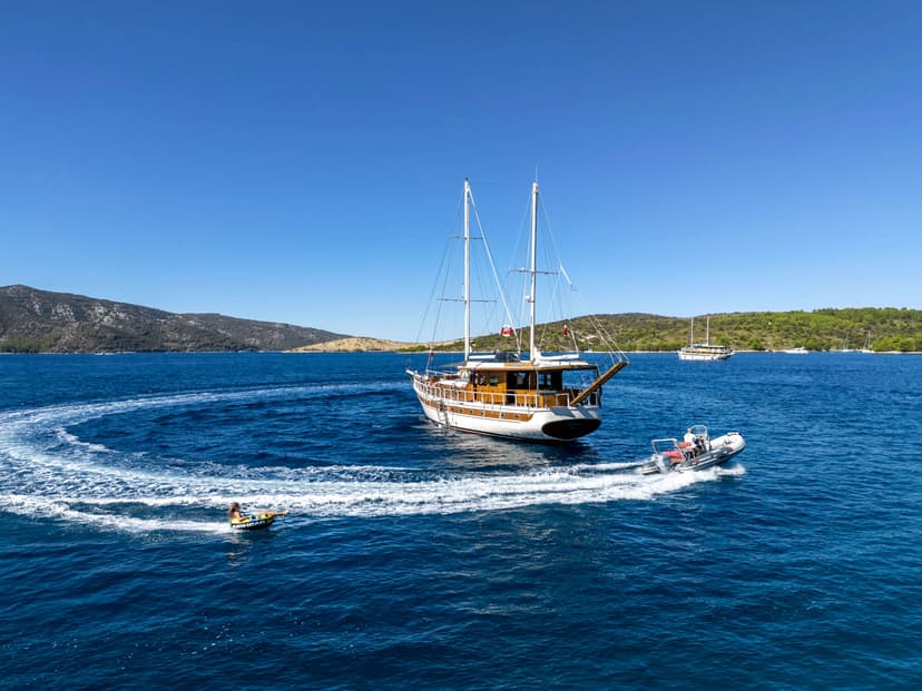 Towed inflatable tubing behind a speedboat near a large wooden sailboat and green hills.