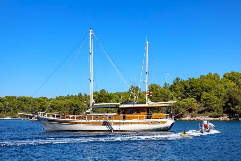 Large wooden sailboat moving on blue water near forested coastline with dinghy towing person.