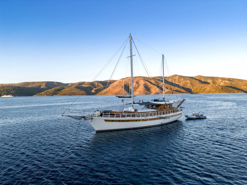 Large wooden sailboat anchored in deep blue water near arid, sunlit hills under clear sky.