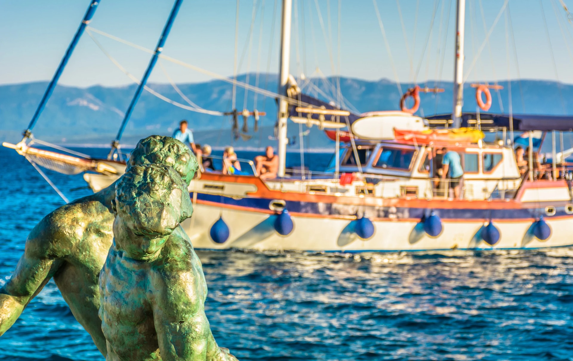 Daily trip on Island of Brac / .Sculpture of fisherman in foreground with touristic boat in background in Island of Brac, Bol town, Croatia.