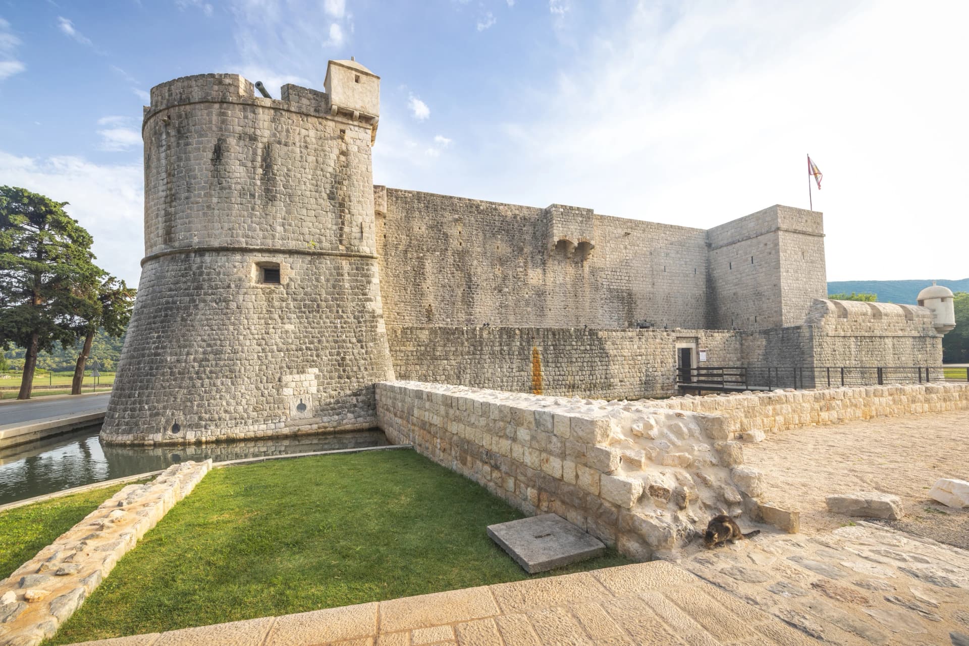 Ston, historic town in Peljesac peninsula in Croatia, Europe. Ancient stone castle with a round tower, surrounded by a moat and grassy area under a bright blue sky with scattered clouds.