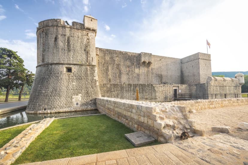 Ston, historic town in Peljesac peninsula in Croatia, Europe. Ancient stone castle with a round tower, surrounded by a moat and grassy area under a bright blue sky with scattered clouds.