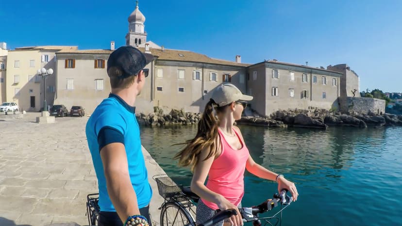 A couple in full caps standing on their bikes, taking a selfie and admiring the view on the Mediterranean Sea. Behind them there is an old city center of Krk. Active traveling. Clear and sunny weather