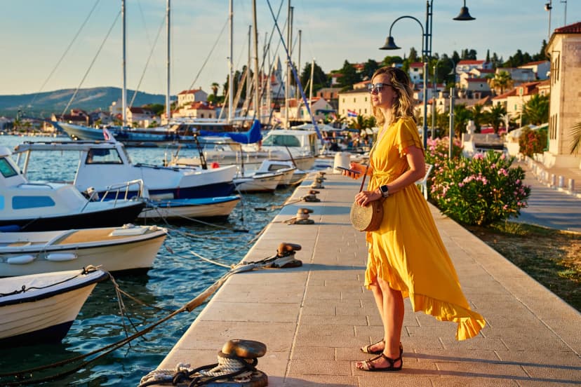 Woman is walking alone on sunny promenade with palm trees and flowers. Female tourist strolls along marina at sunset in coastal town